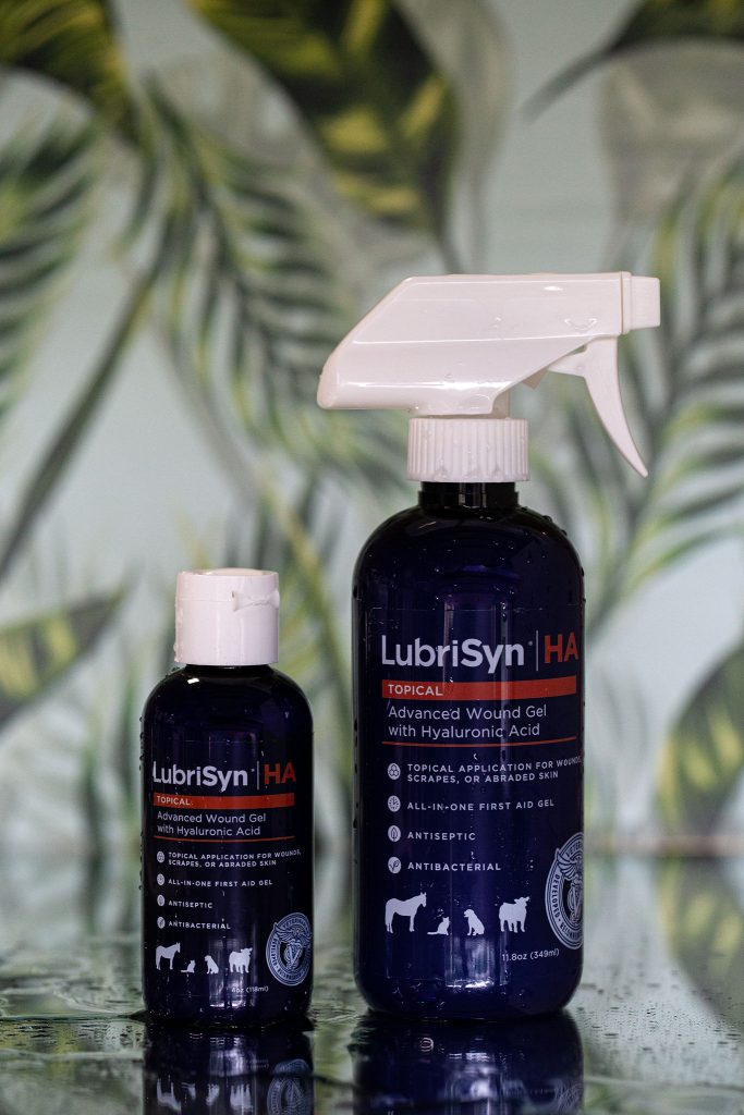 A small bottle of LubriSynHA Topical Wound Gel next to a large bottle of LubriSynHA Topical Wound Gel in front of a leafy background.
